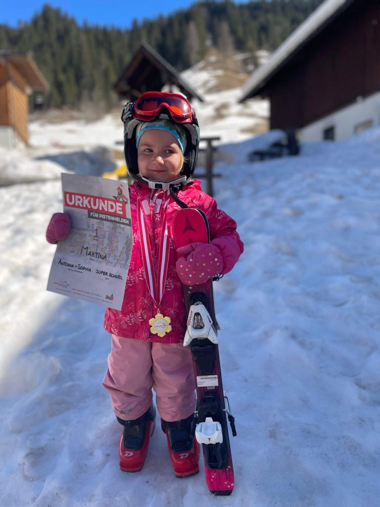 Ein kleines Mädchen hält eine Urkunde und steht im Schnee mit Skiausrüstung.