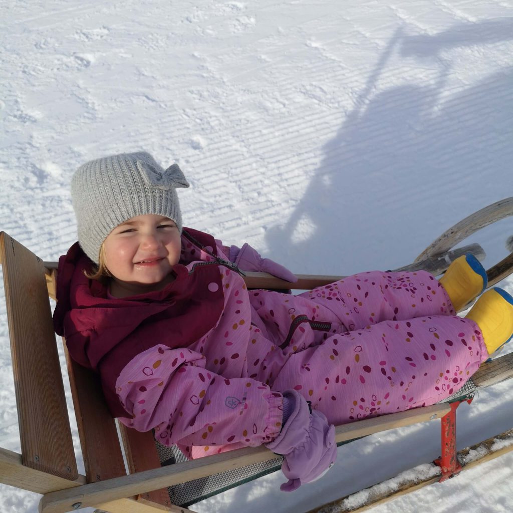 Kind in rosa Schneekleidung sitzt auf einem Schlitten im Schnee, strahlend und glücklich.
